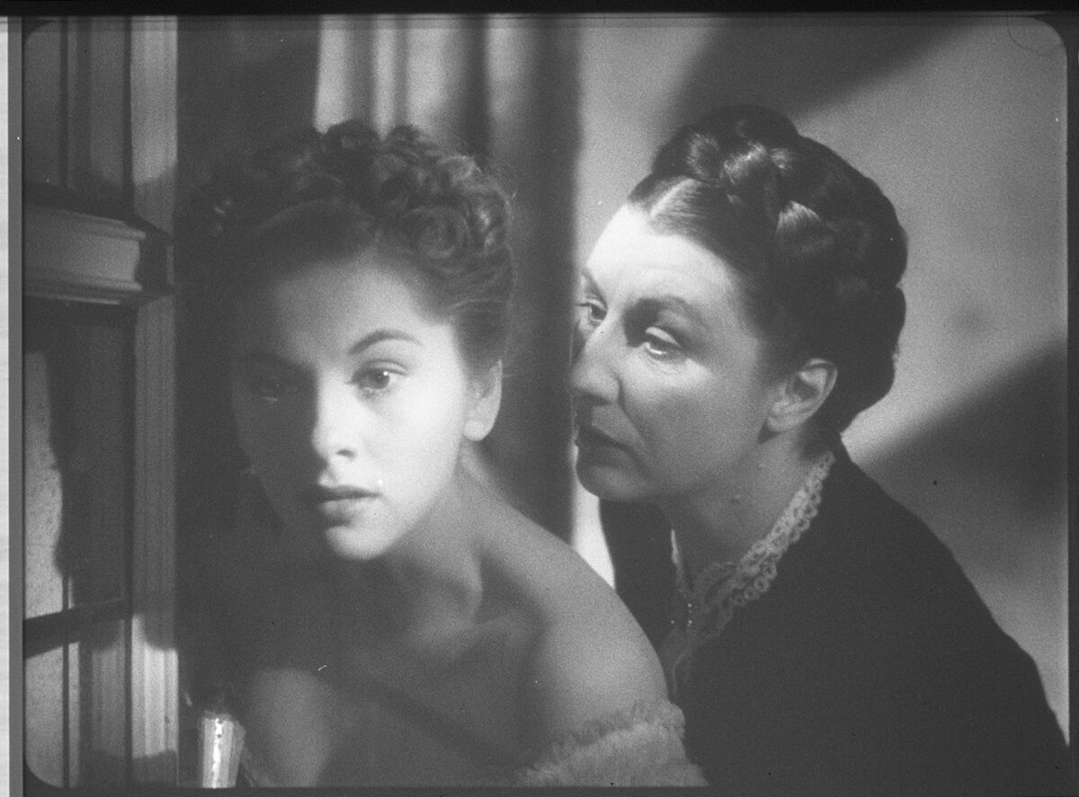 A young woman stands near a window, an older woman behind her leaning sinisterly toward her