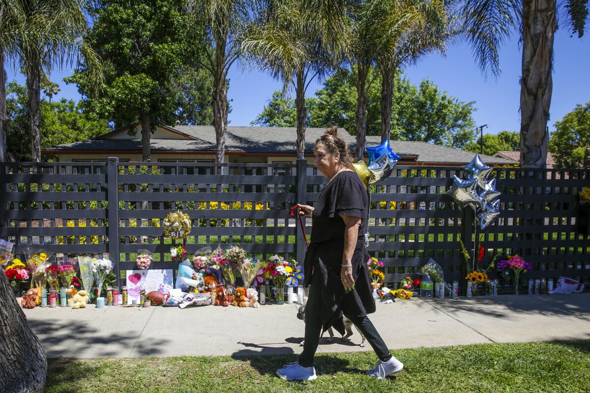 A woman walks a dog past a memorial of flowers, balloons, candles and stuffed toys.