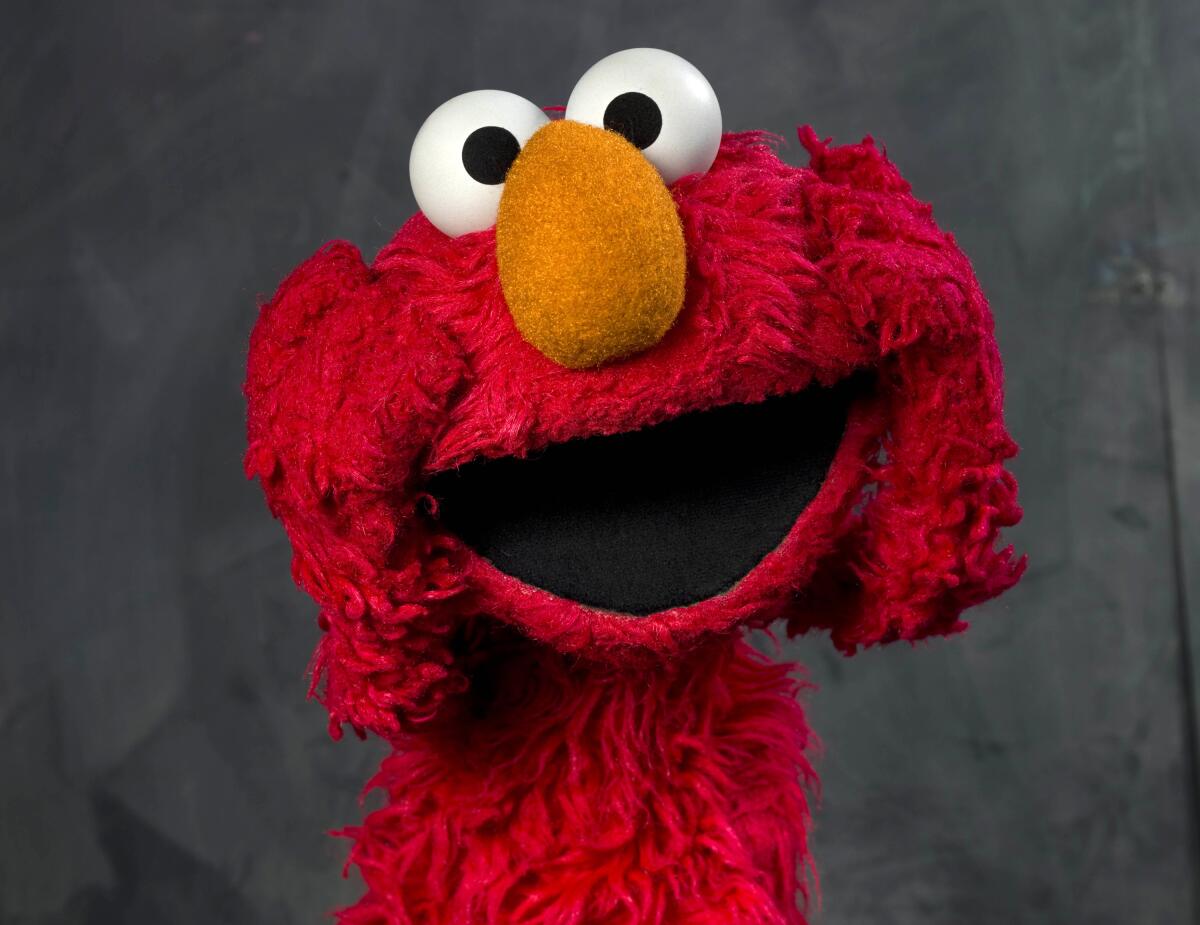 The red muppet named Elmo holds his furry hands to his face while opening his mouth