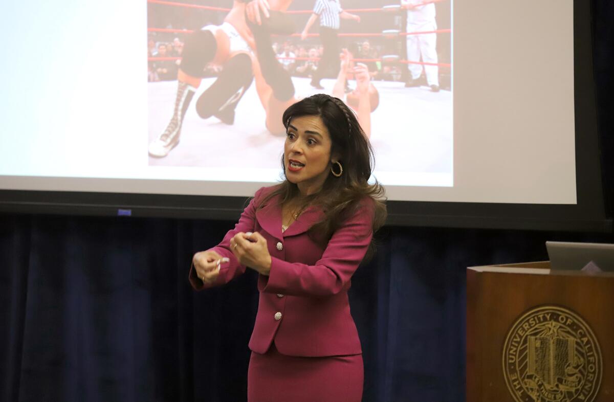 A woman in a red suit speaks in front of a projection screen showing a wrestling match