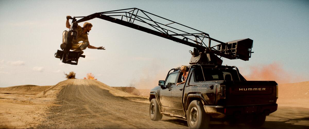 A man seated on a movie crane speaks to a woman in a car.