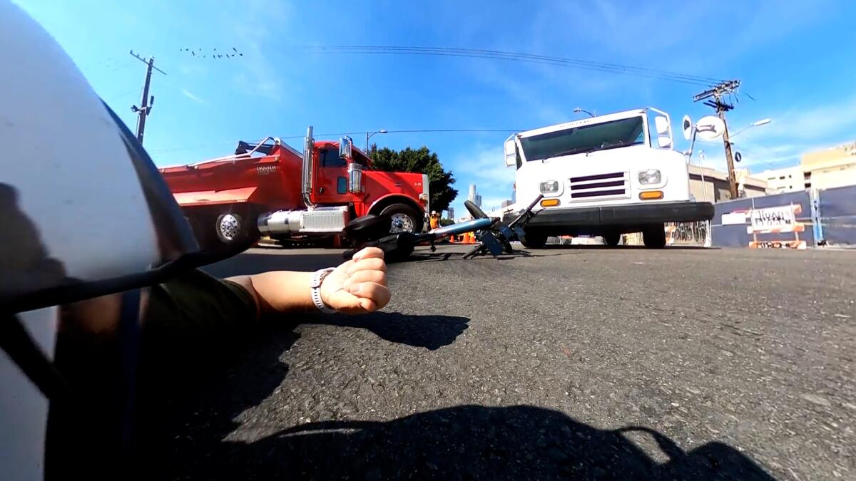 A person lies in the street near a bicycle strewn between a red big rig and a USPS truck.
