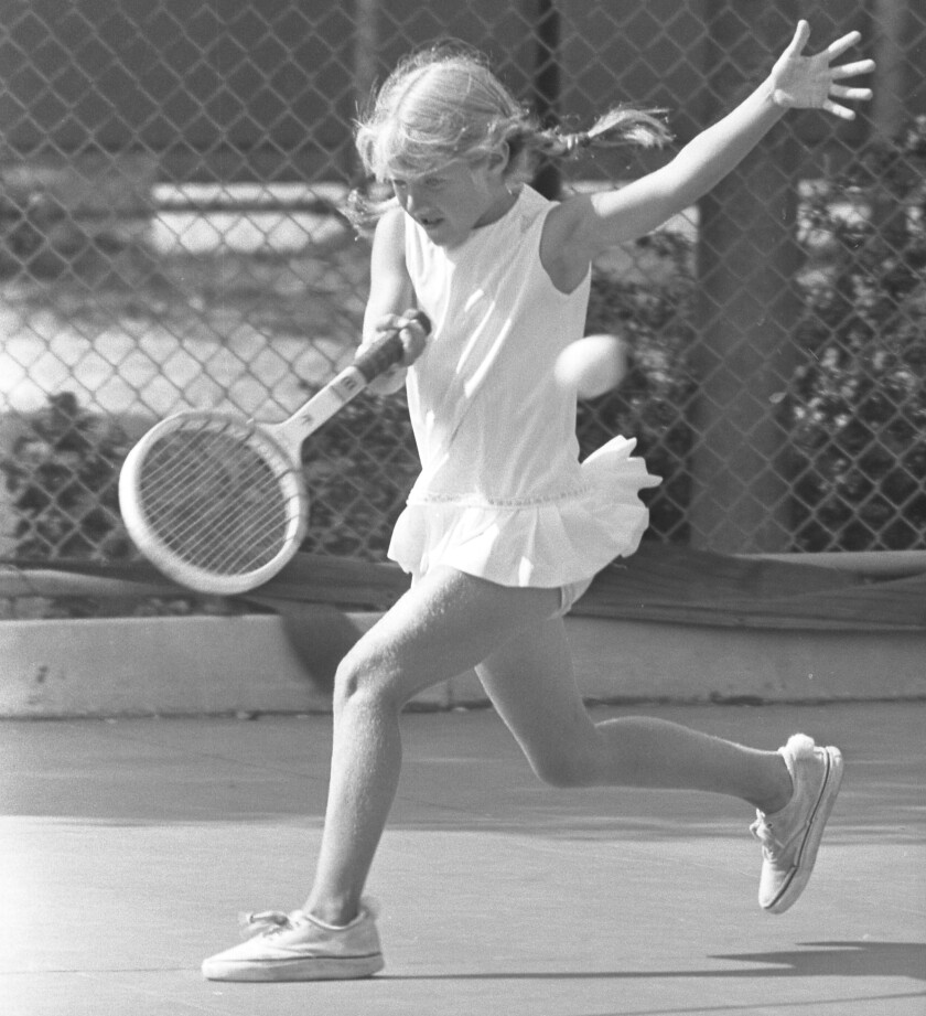 A young girl in a white tennis outfit and braids swings a wood racket as she returns a ball on court.