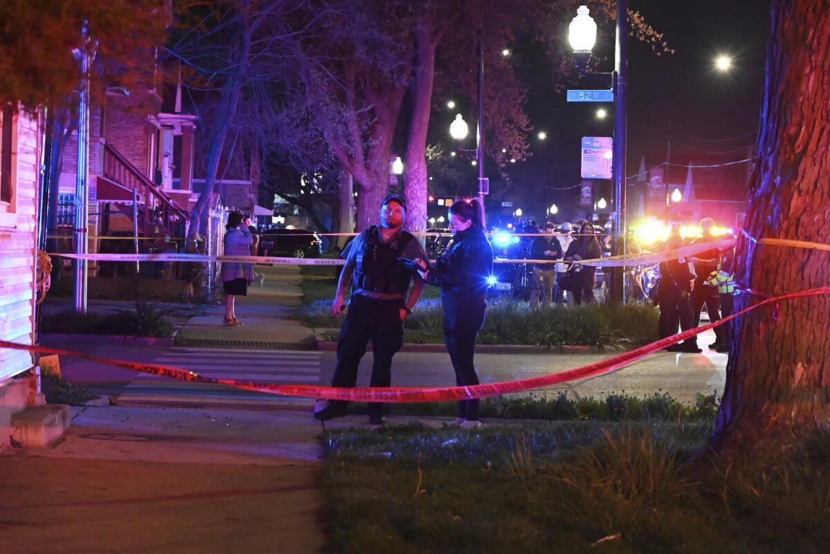 Police tape crosses a sidewalk at night as an officer talks to a person standing nearby.