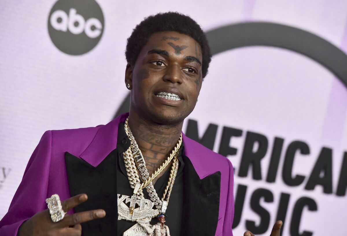 A man in a purple tux with black trim wears several heavy gold and diamond chains and smiles to show off his grills.
