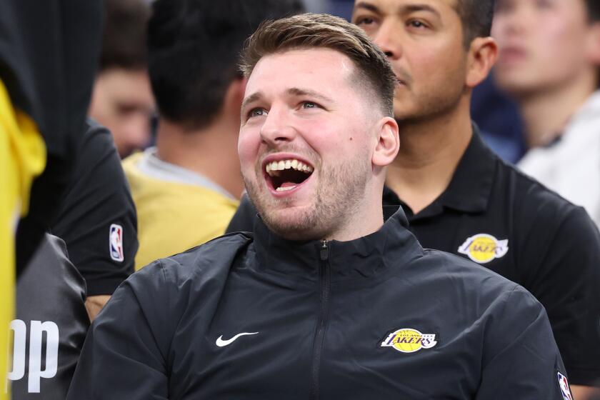 Luka Doncic laughs while sitting on the Lakers bench in street clothes during a timeout