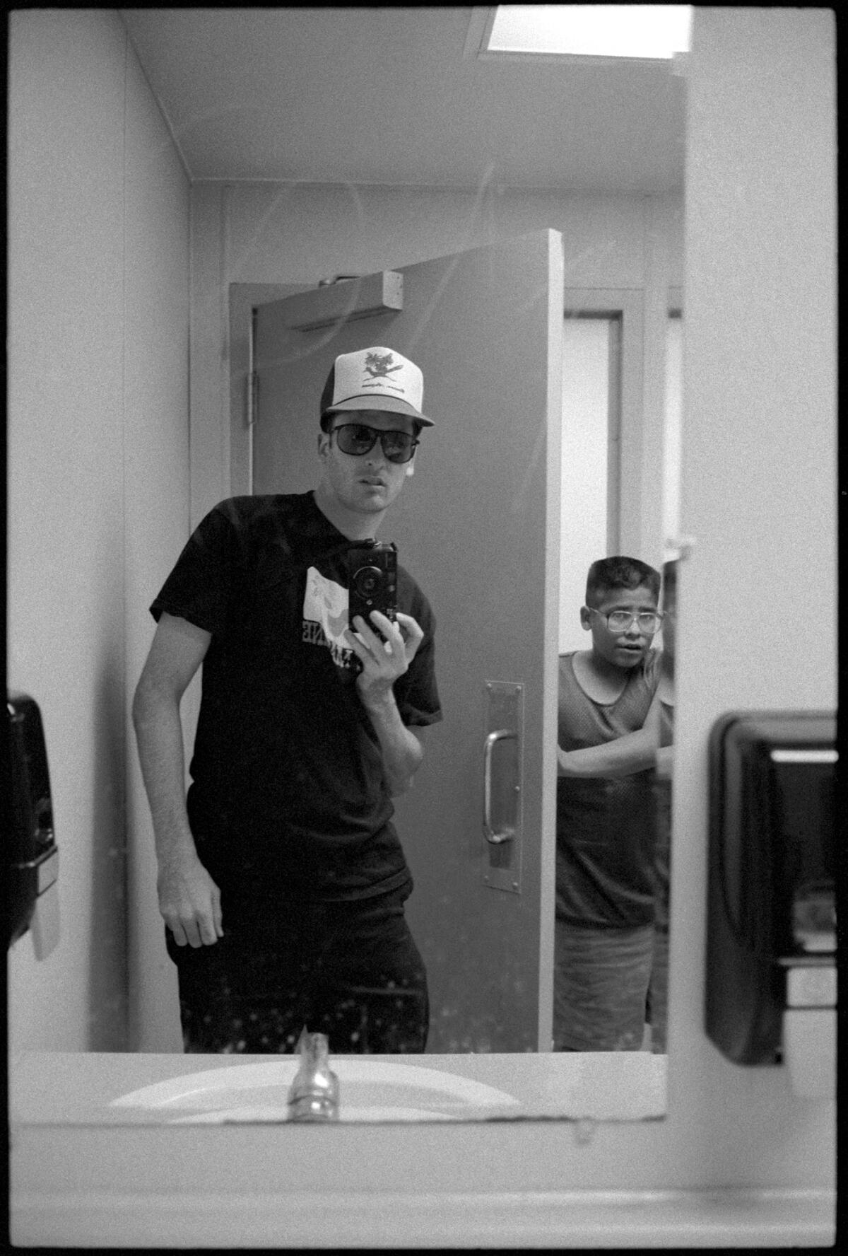 A young man in a baseball cap takes a selfie in a bathroom mirror as another person walks in.