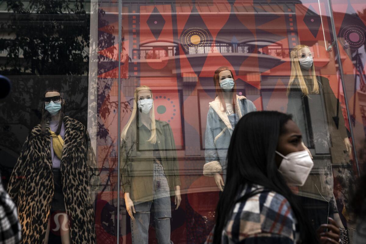 A masked pedestrian walks past mannequins also wearing face masks in a store window.
