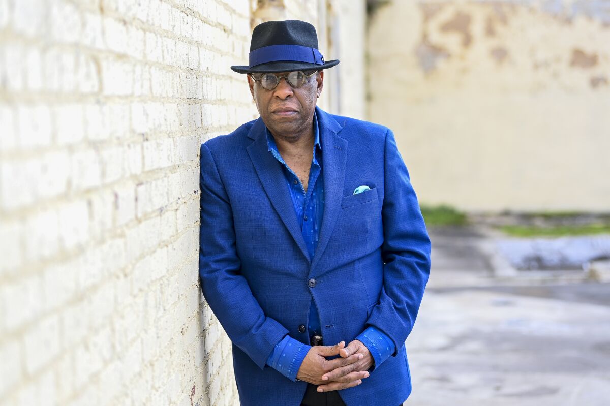 A man in a blue suit and fedora leans against a brick wall.