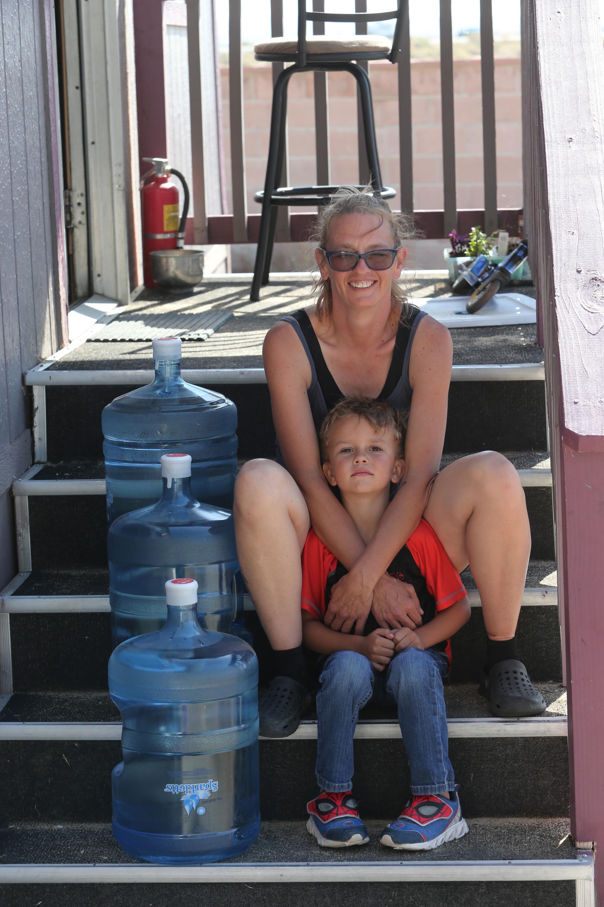 A woman and a young boy sit on a porch beside several five-gallon bottles of water.