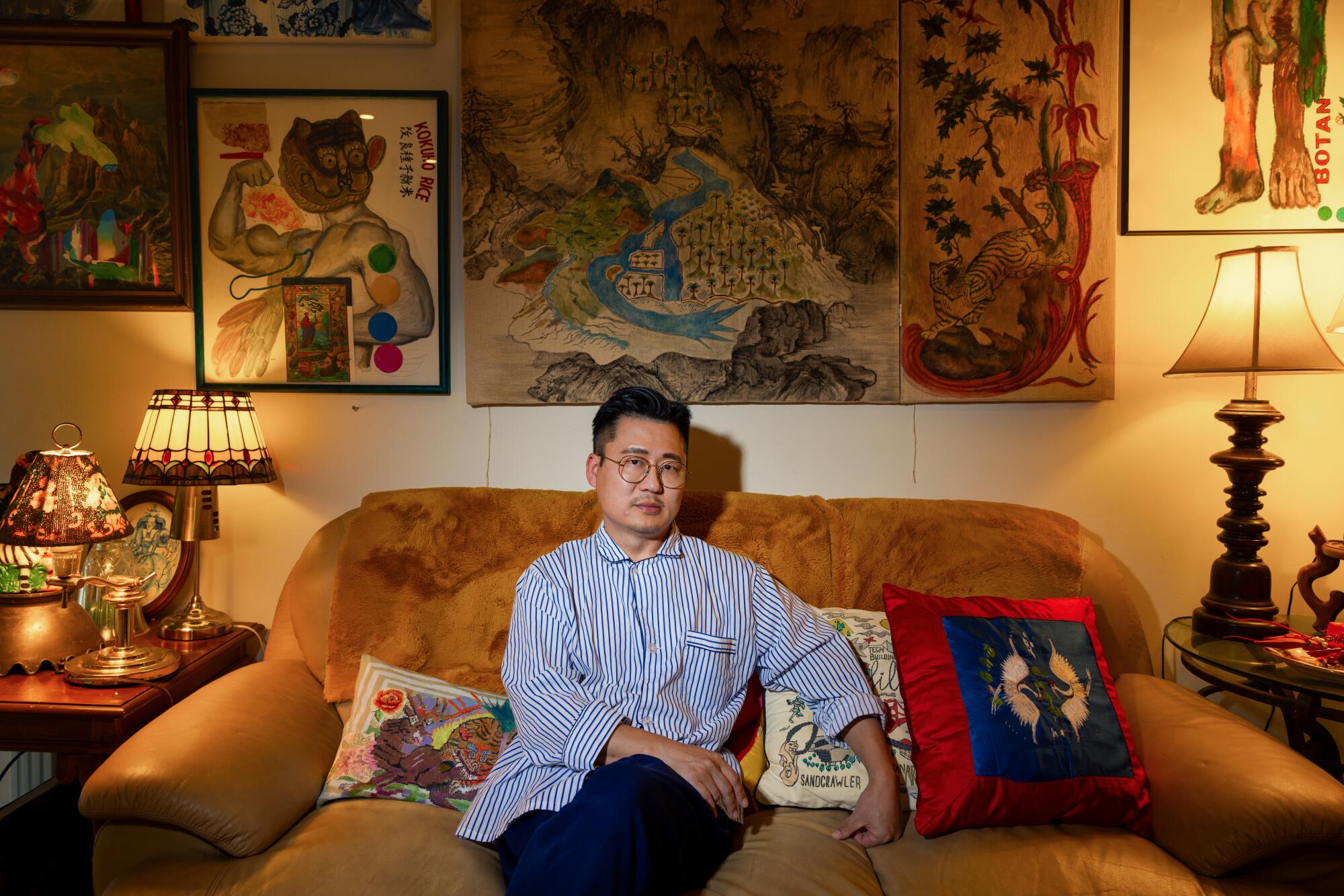 O artista Ken Gun Min está sentado em um sofá cercado por suas obras de arte