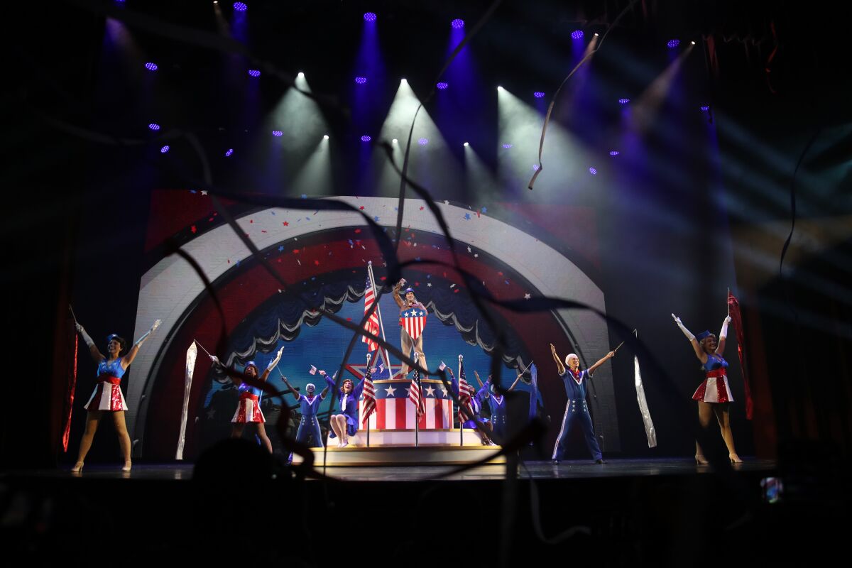 Actors on a stage wear red white and blue outfits and stand on red white and blue set pieces.
