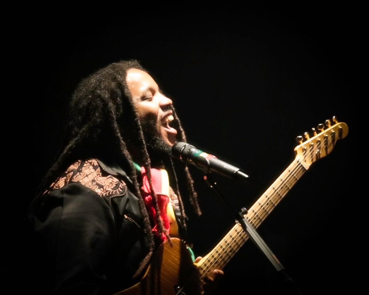 A man with dreadlocks plays the guitar and sings into a microphone