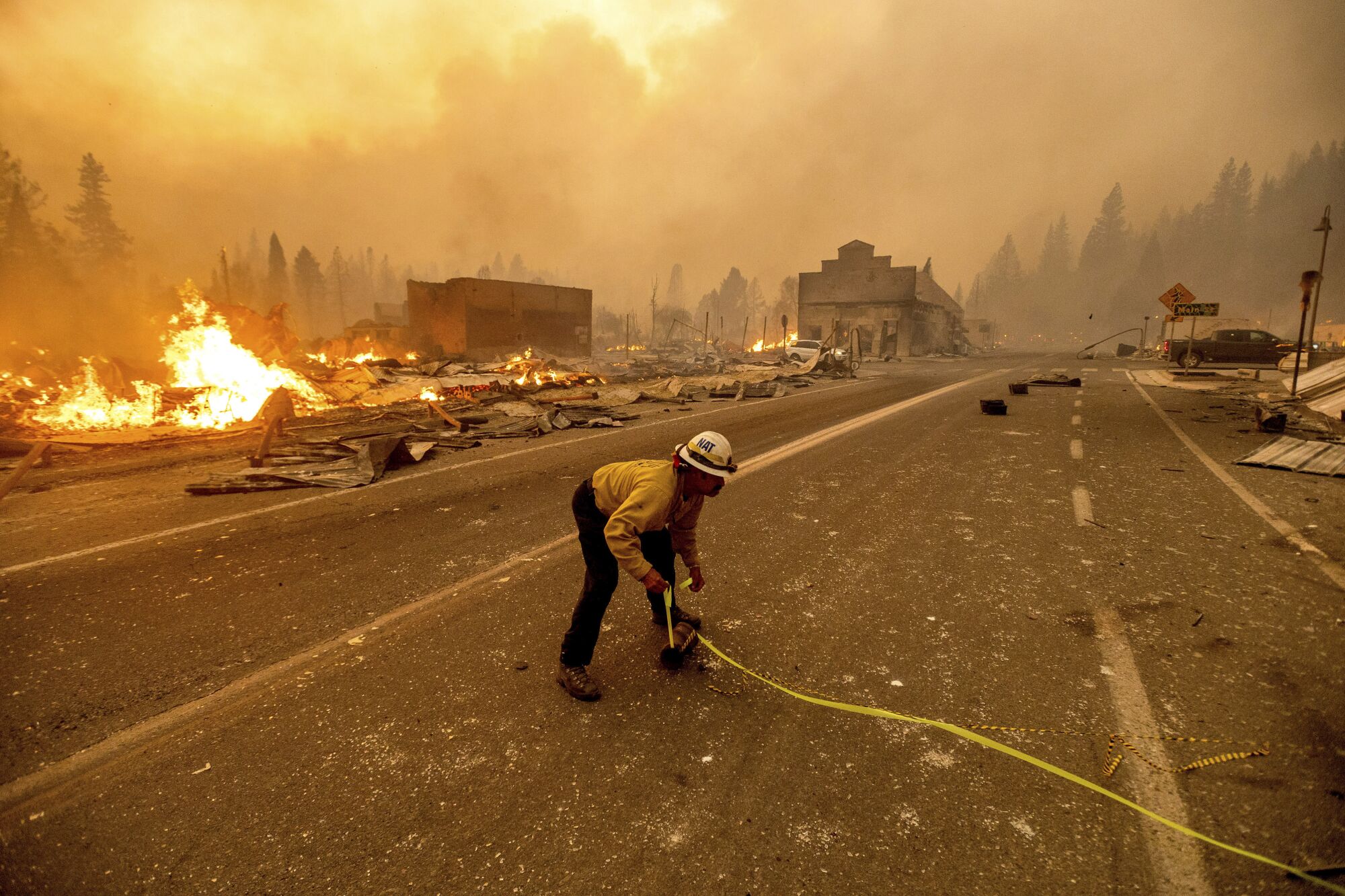 A man bends as he uses tape to mark a roadway. In the background, fire burns and ruined buildings are shrouded in smoke.