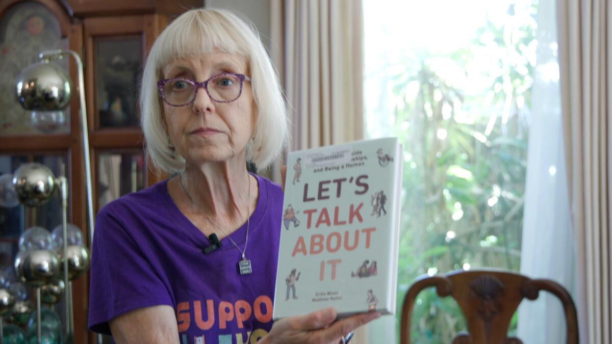 A seated woman with white hair holds up the book "Let's Talk About It."