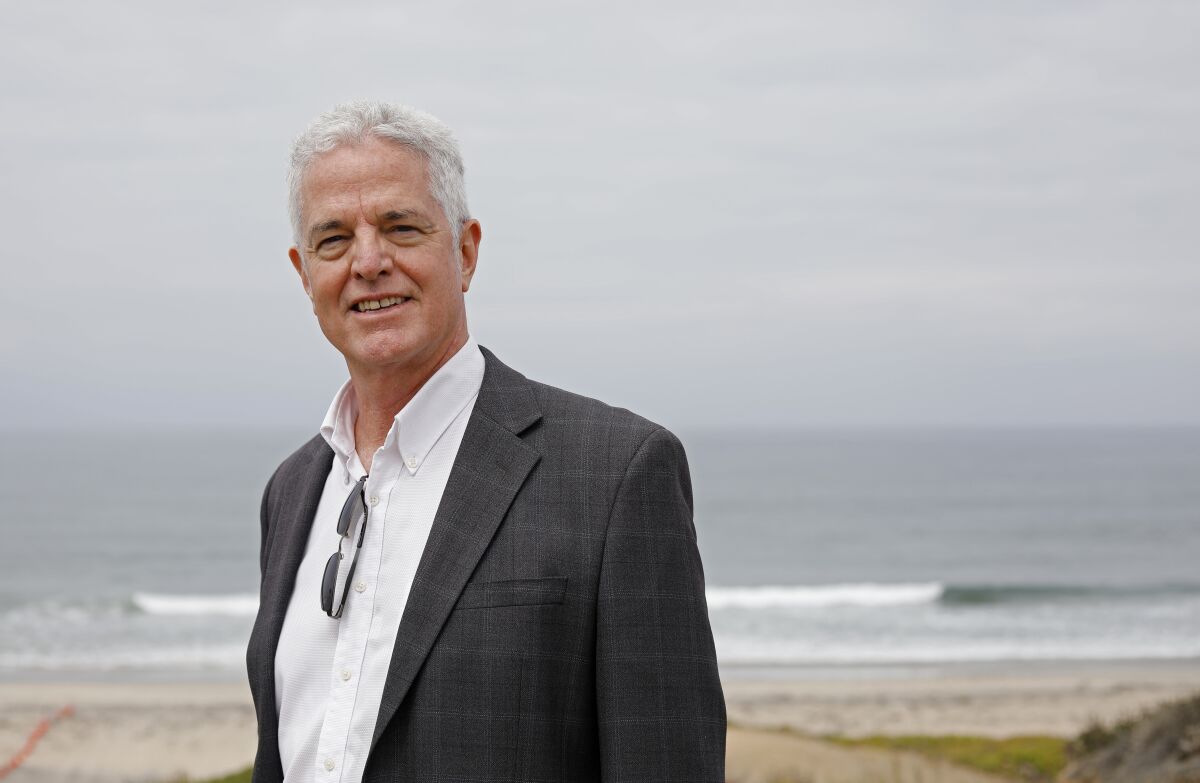 Author John Fanestil is photographed against the Pacific Ocean, wearing a black jacket and white button-down shirt. 