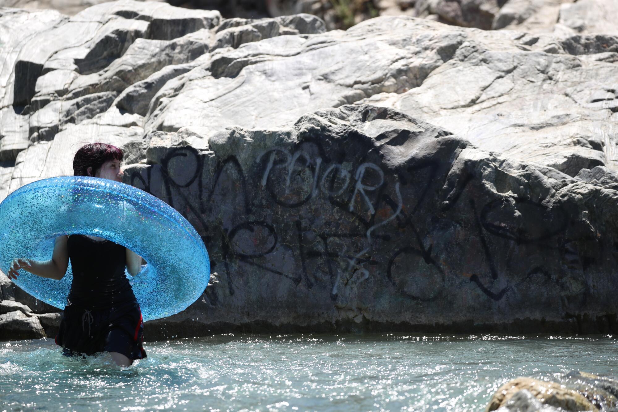 A woman standing in a river holds a blue innertube while graffiti covers rocks in the background. 