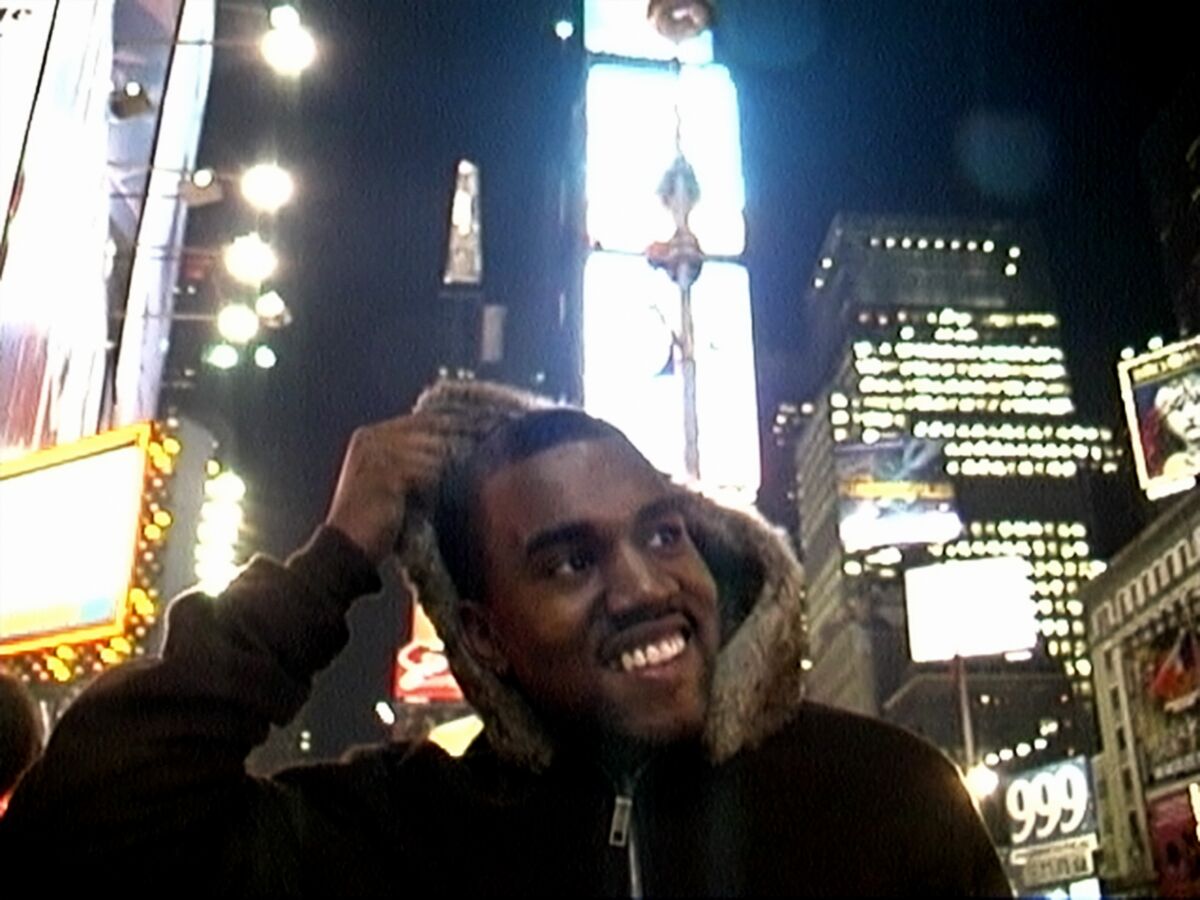Kanye West holds his hand above his head in front of brightly lit buildings