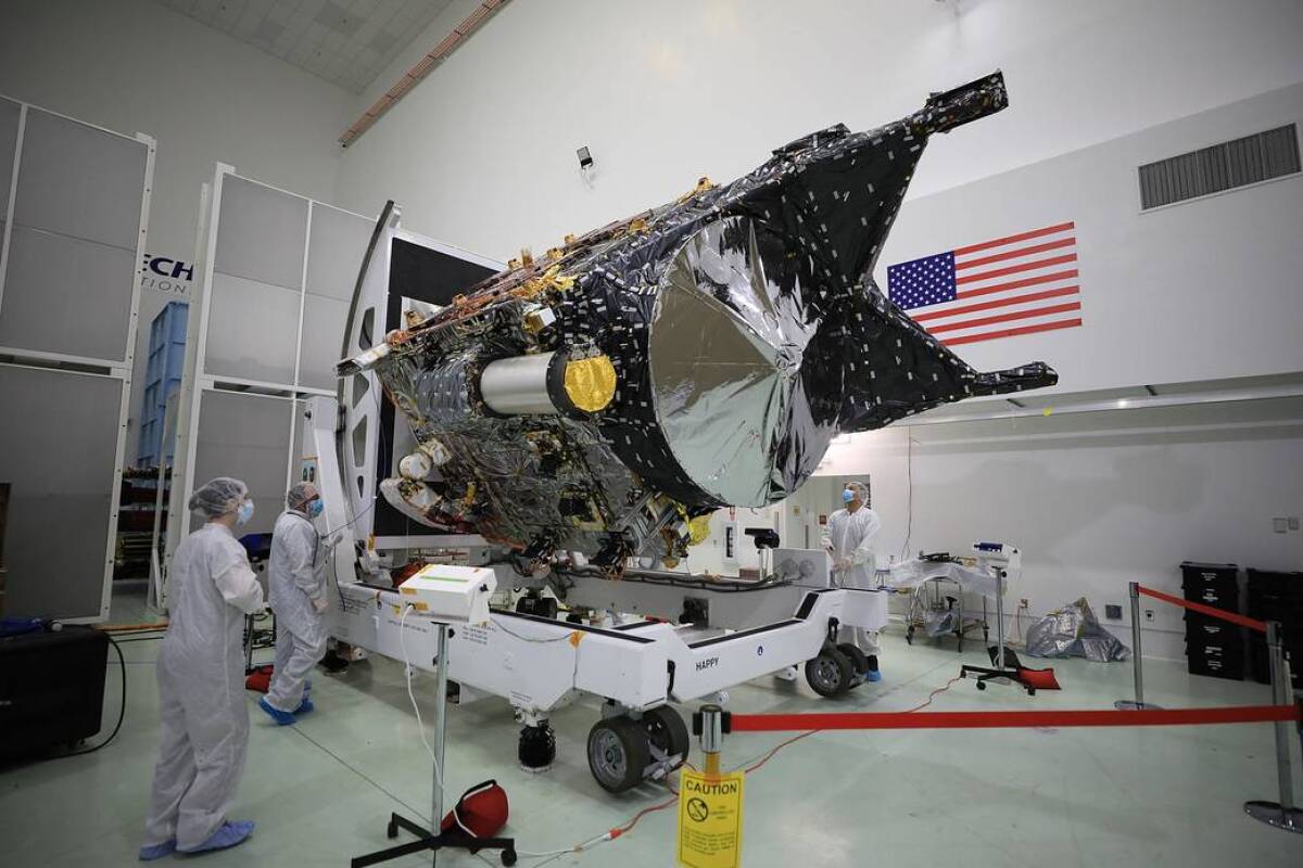 Workers in white protective suits look at a spacecraft in a white room with a U.S. flag on one wall