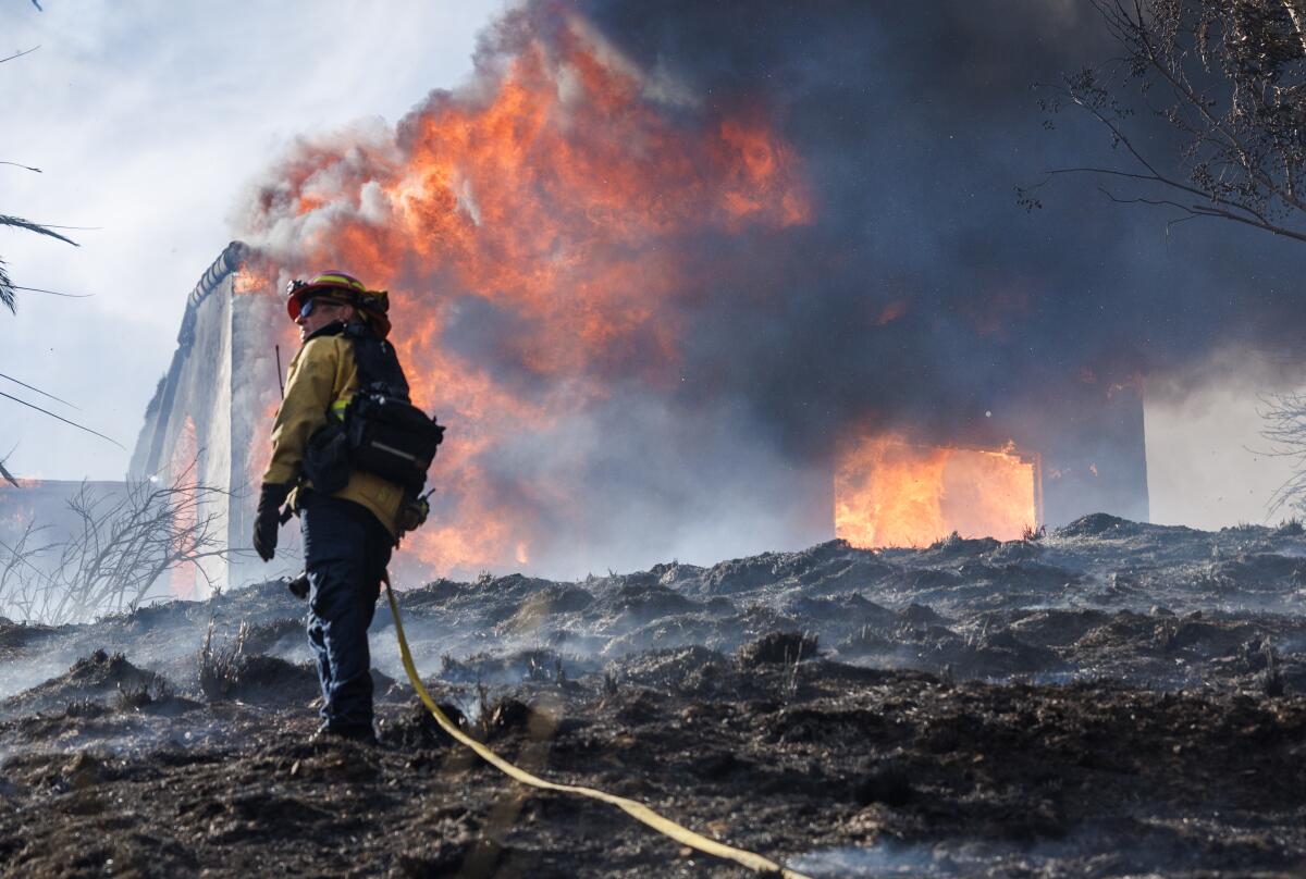 A firefighter standing on smoldering ground in front of a burning house