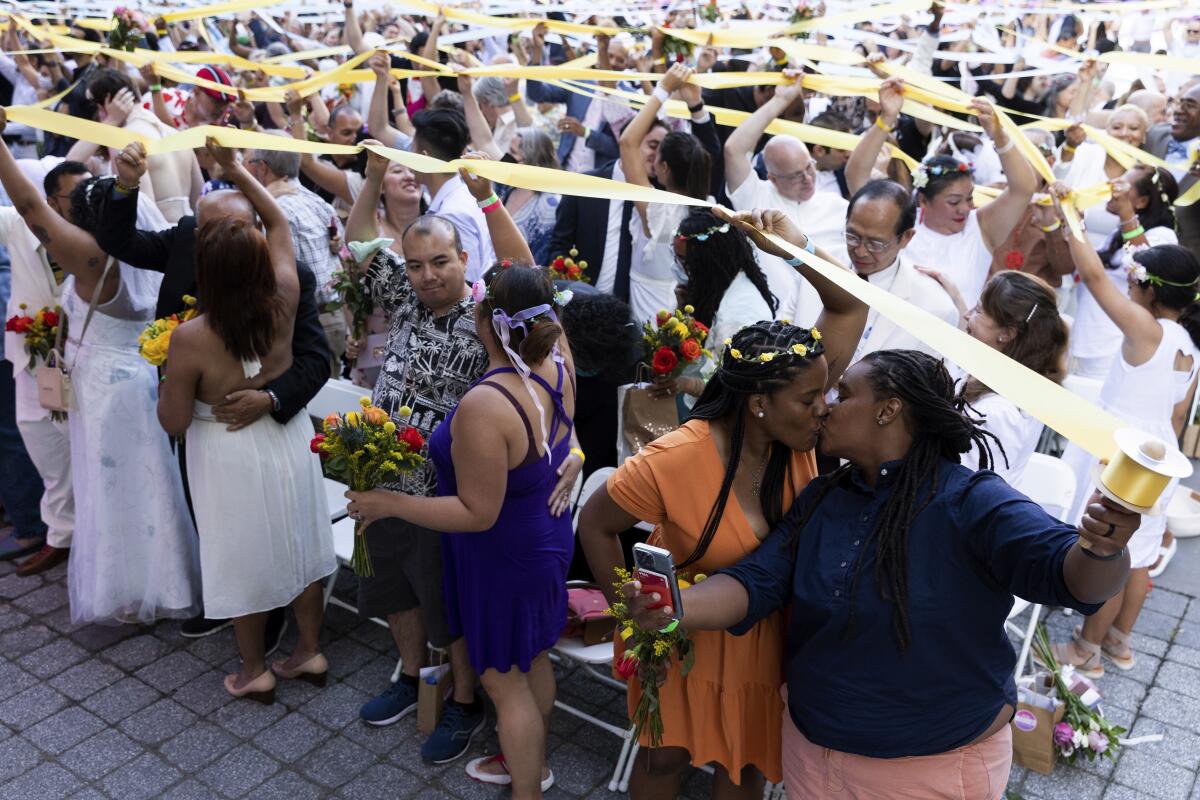 People, some in white dresses, stand in pairs, some kissing and some holding up yellow streamers.