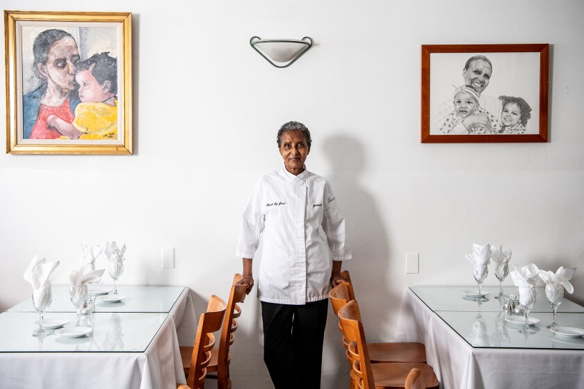 A woman chef standing up against a wall in between her restaurant tables.