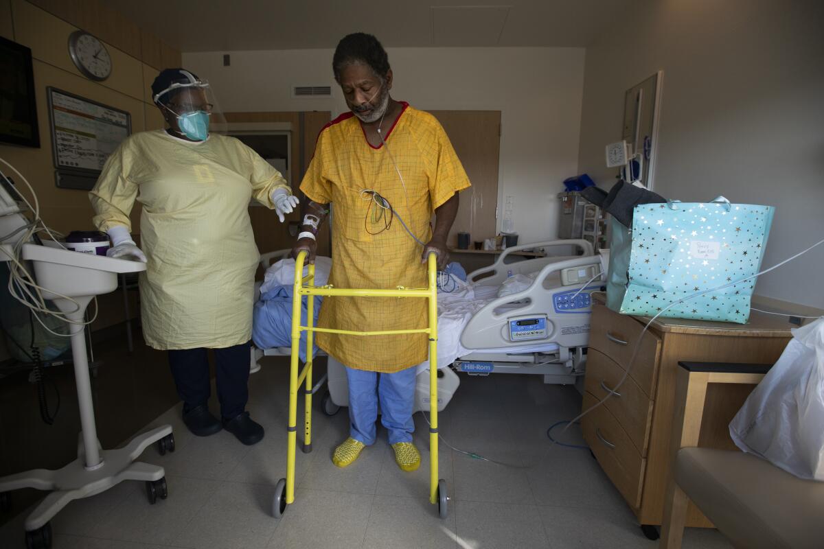 A hospital patient stands next to his bed with the use of a walker as a nurse in protective gear watches over him