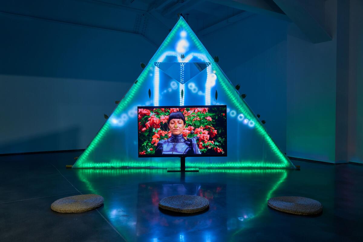 A video monitor displaying a woman in futuristic black attire sits on a glossy floor with a backdrop of neon light.