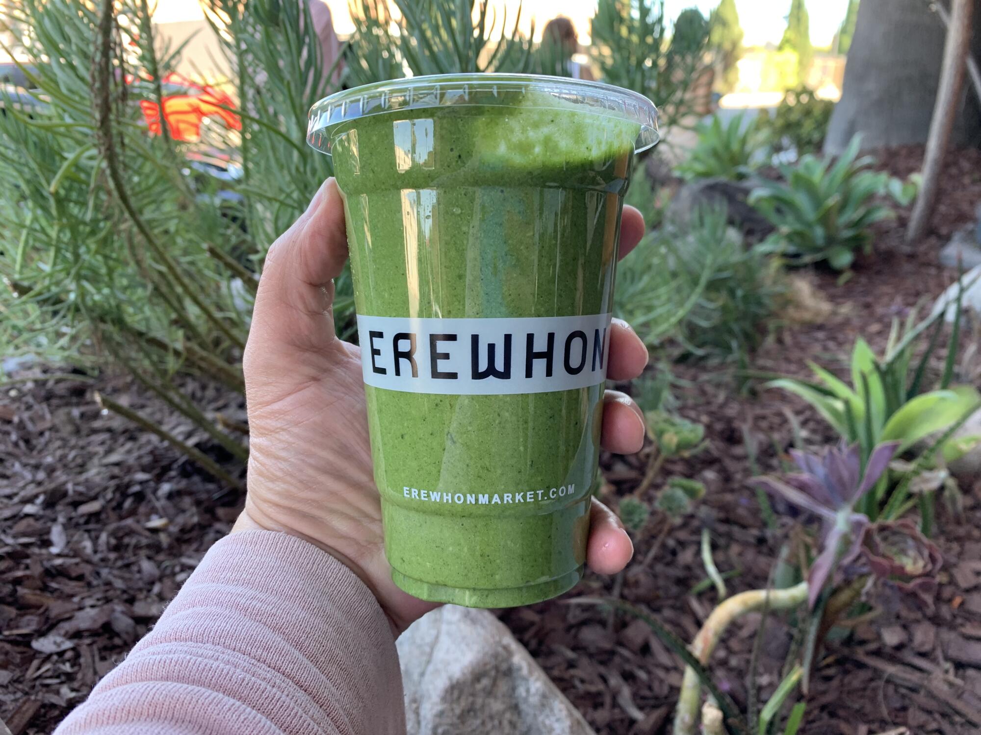 A hand holds a green smoothie in a plastic cup that says Erewhon.