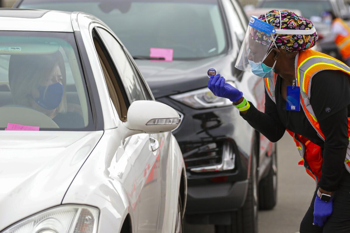 A nurse offers "I Got My Covid-19 Vaccine!" sticker to person in a vehicle.