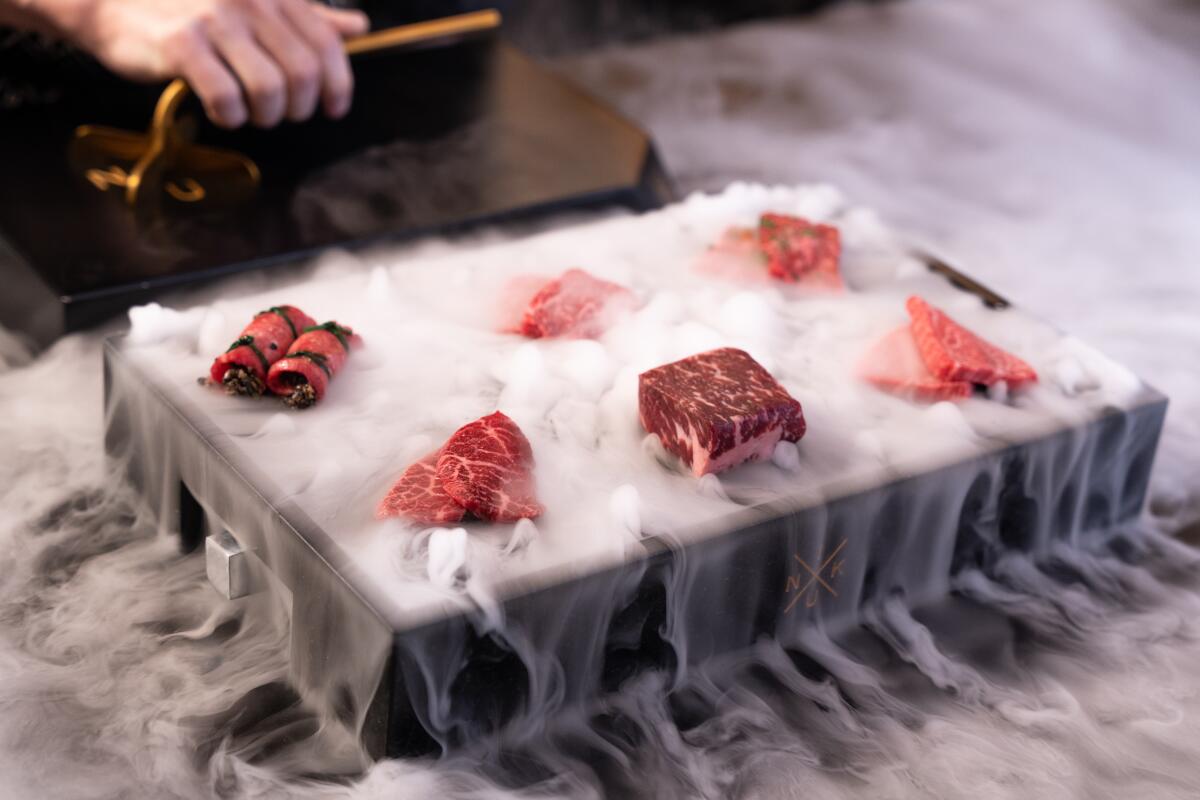 Six small cuts of raw meat sit on a tray. Smoke or dry ice billows around them.