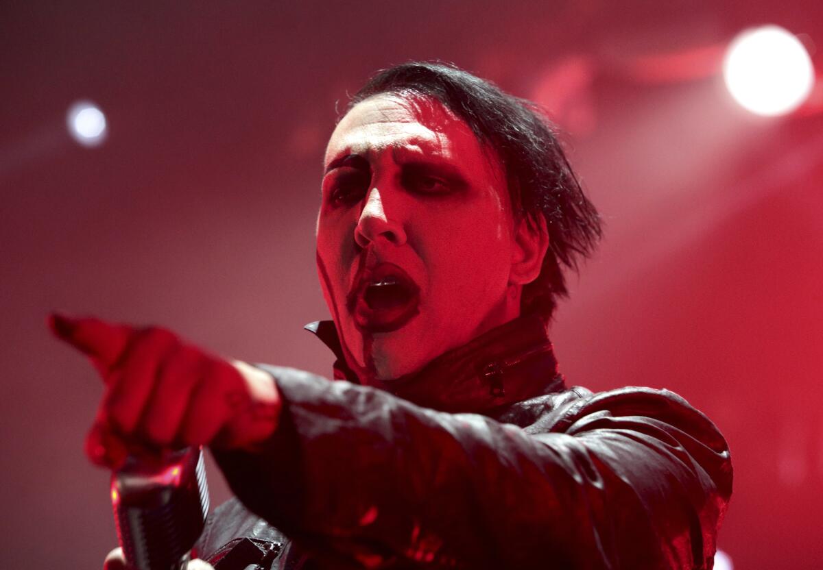 A man in Goth makeup and a leather jacket points forward while performing on a stage filled with red light