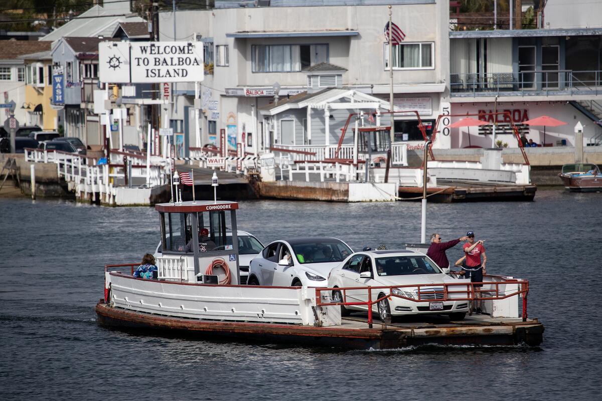 A small ferry boat carrying three vehicles pulls away from a landing. A sign reads "Welcome to Balboa Island."