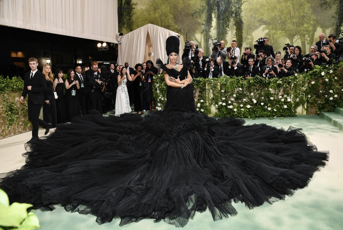 Cardi B with arms crossed wearing a large black turban and a black gown with its massive skirt fanned out on a green carpet