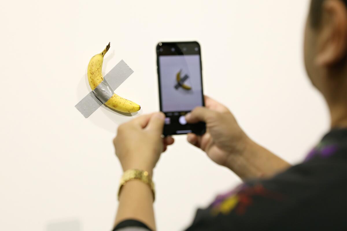 A man takes a cellphone photo of a banana taped to a wall.