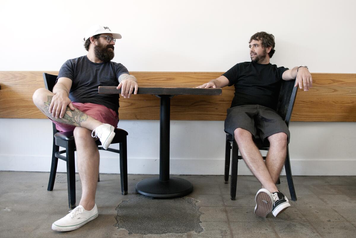 Two men wearing shorts seated on either side of a small table talking to each other