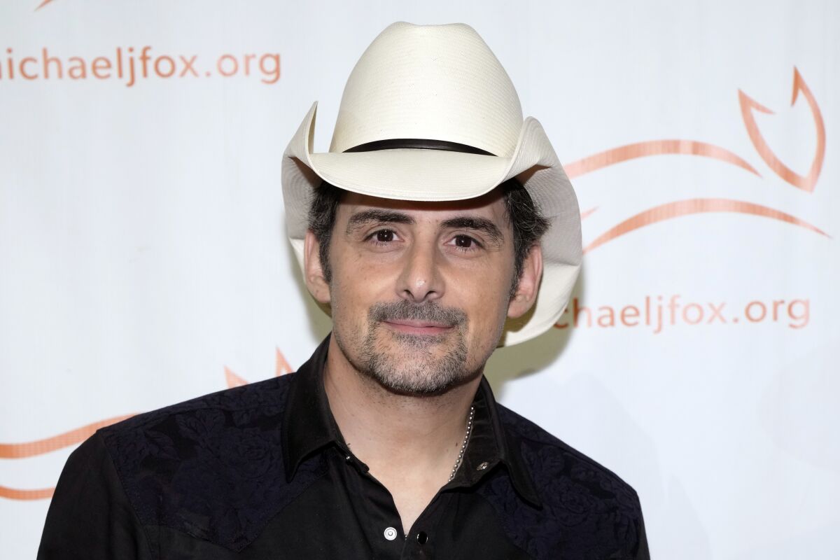 A man wearing a white cowboy hat and a black shirt poses in front of a backdrop