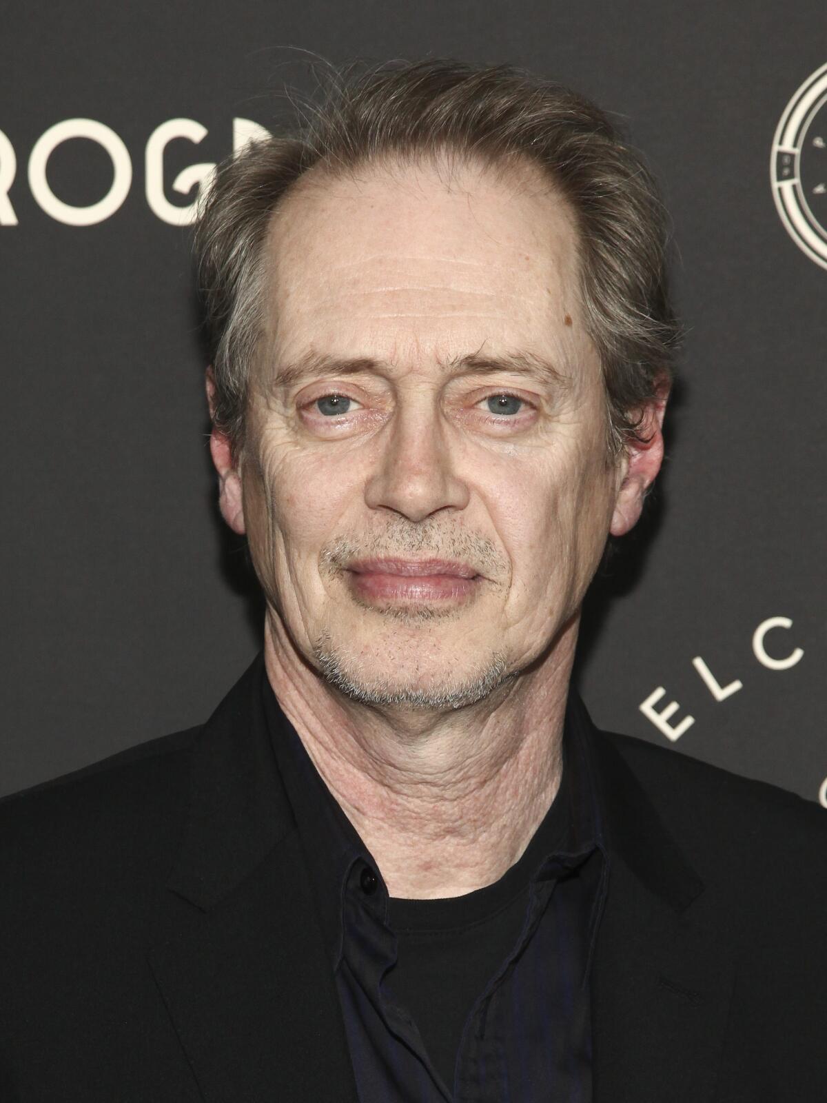 Steve Buscemi wears a black shirt and stands in front of a black backdrop with white writing