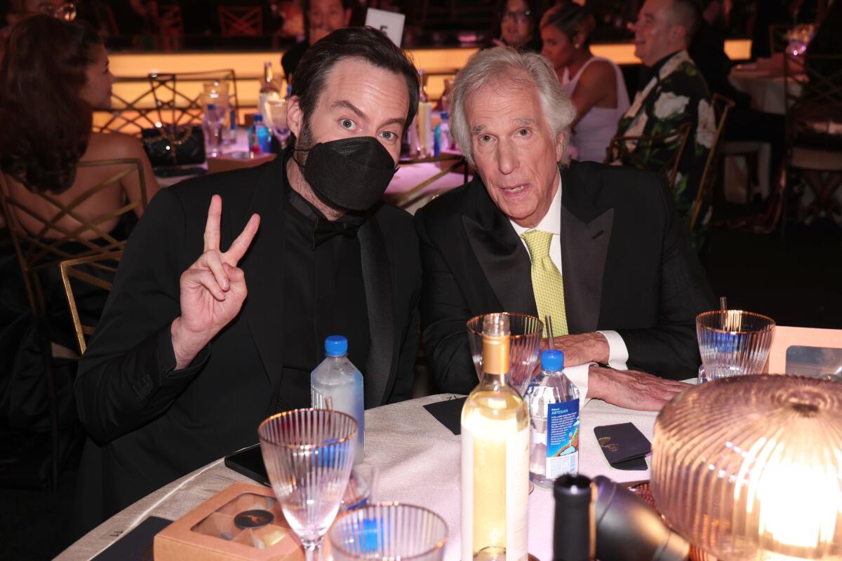 A man wearing a mask flashes a peace sign while seated next to another man at a table.