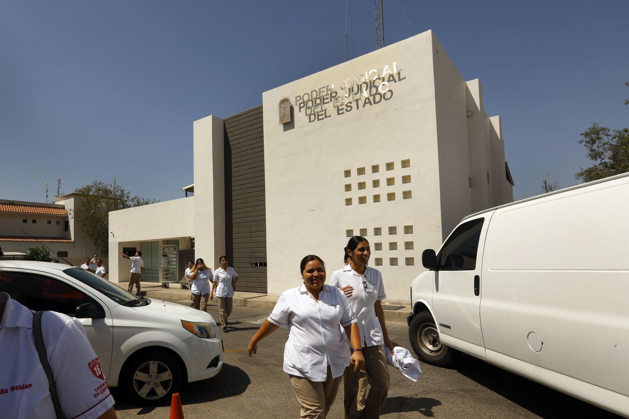 Dos mujeres con uniforme blanco y pantalones caqui caminan cerca de un edificio de color beige y autos