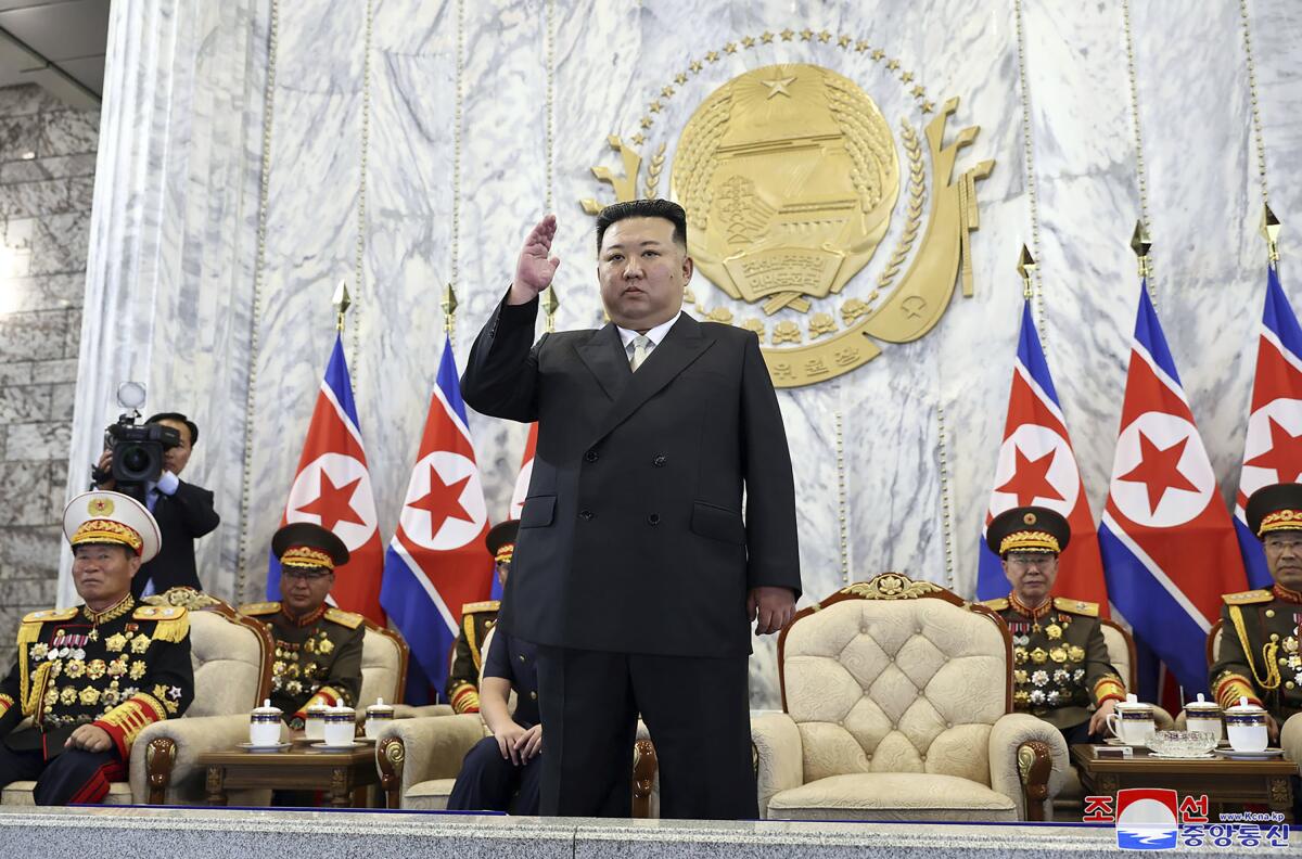 Kim Jong Un stands with raised hand while men in military uniforms sit in chairs behind him and an insignia is on a wall. 