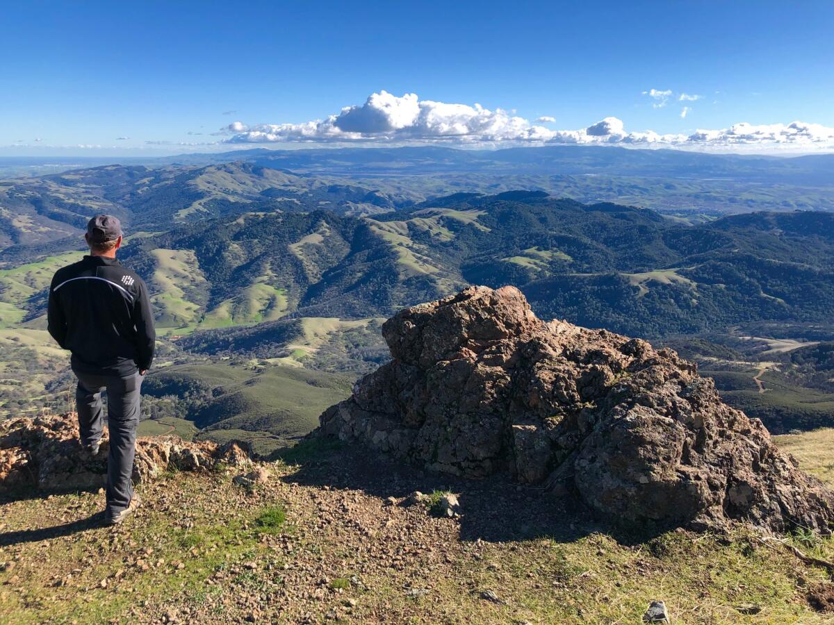 A man on a mountain top looking out at a green valley and blue skies with clouds in the distance.