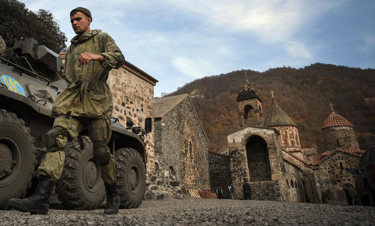 A man in camouflage fatigues walks past an armored personnel carrier parked by a stone monastery.