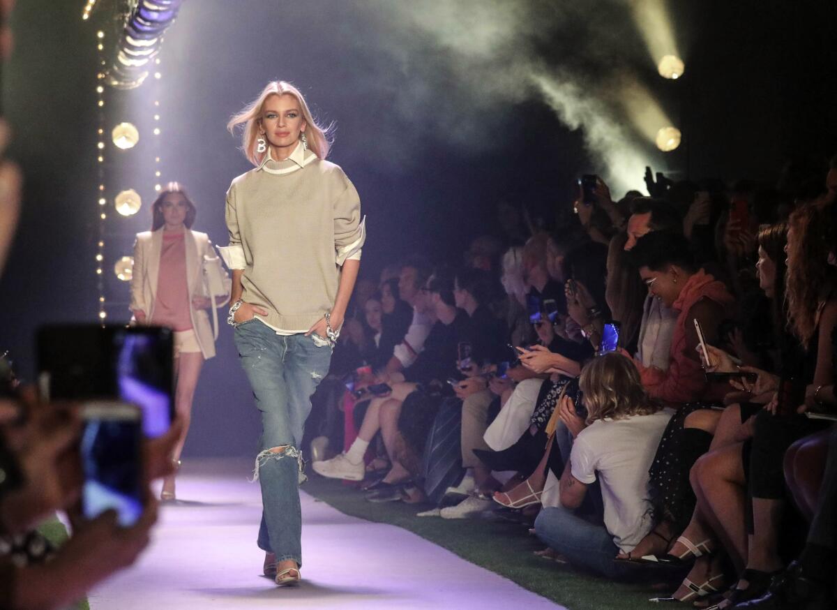A woman walks the runway in a look from Brandon Maxwell: beige sweater over a white collared shirt and blue jeans, ripped at the knee.