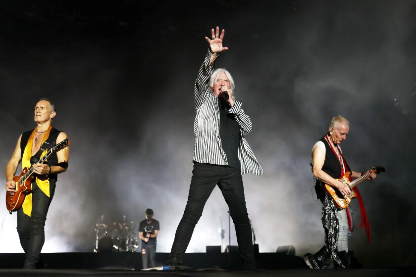 Three men on a stage  -- two men playing guitars on either side of a man singing into a microphone and reaching his hand out