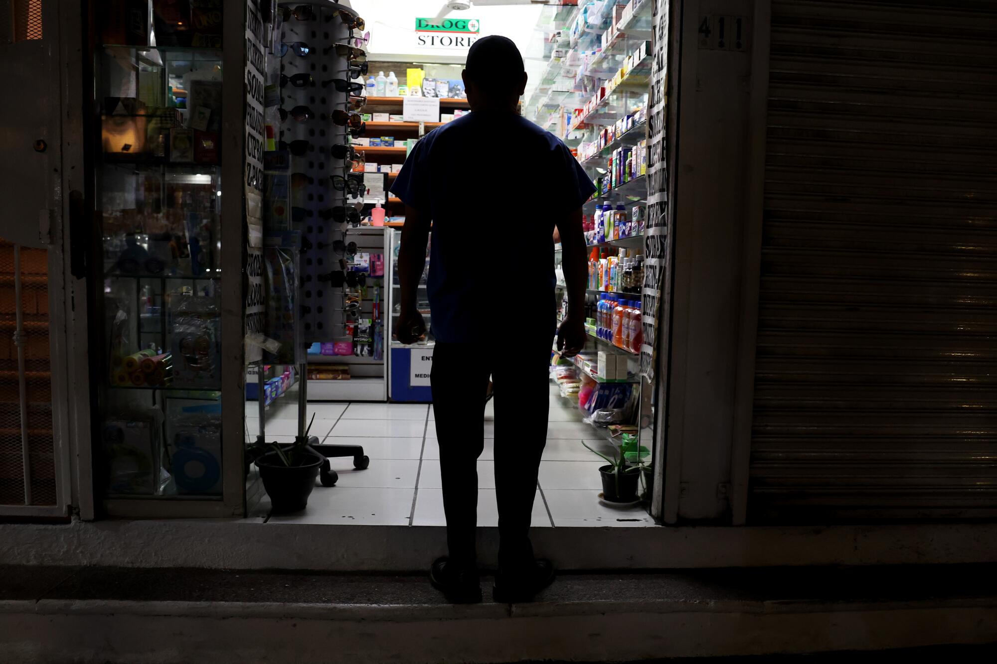 A man appears only as a dark shape in the doorway of a bright drugstore as he enters at night