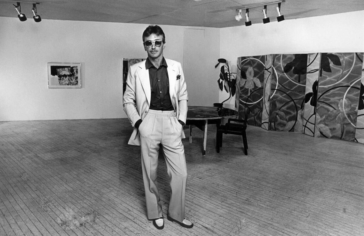 Artist Billy Al Bengston stands in a suit and tinted glasses in a room containing paintings and a table and chair.