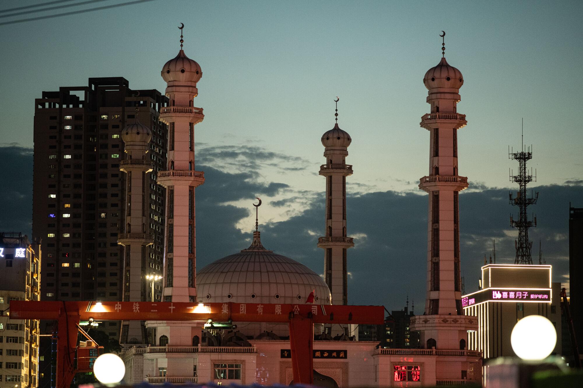 A building with a dome in the center and four minarets on the corners sits next to tall buildings against an evening sky 
