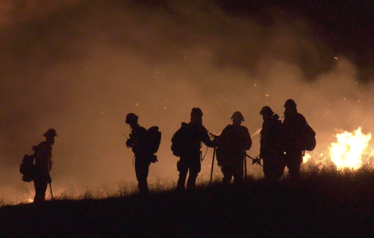 Firefighters are seen in silhouette on a hillside, with smoke and flames in the background.