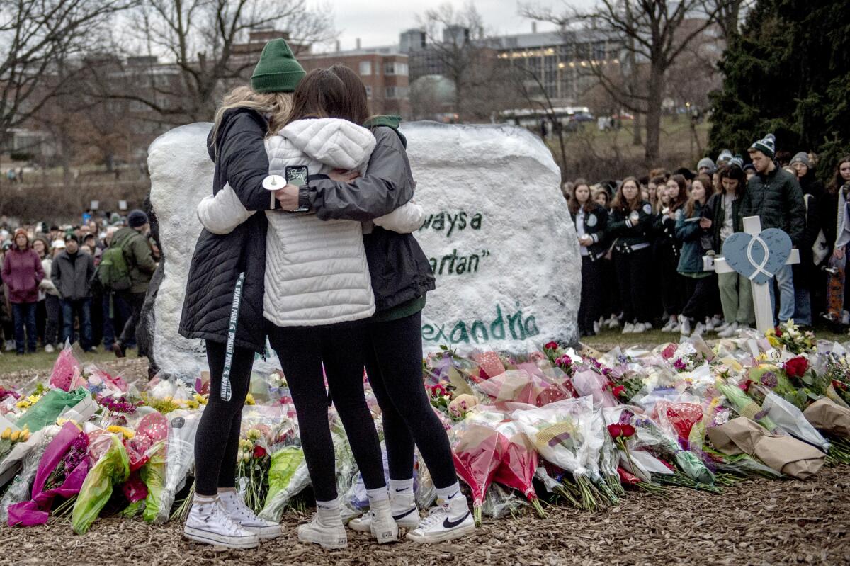 Three students embrace in front of flower bouquets on the ground.